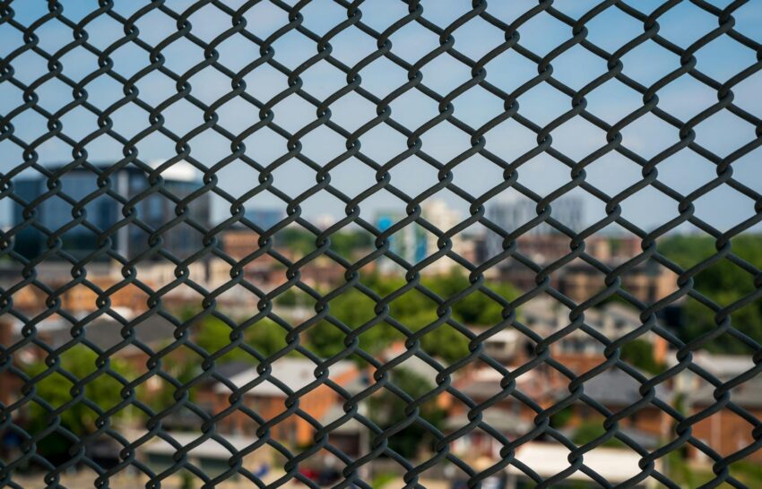 A cityscape blurred behind a metal mesh fence, showcasing urban architecture and greenery.