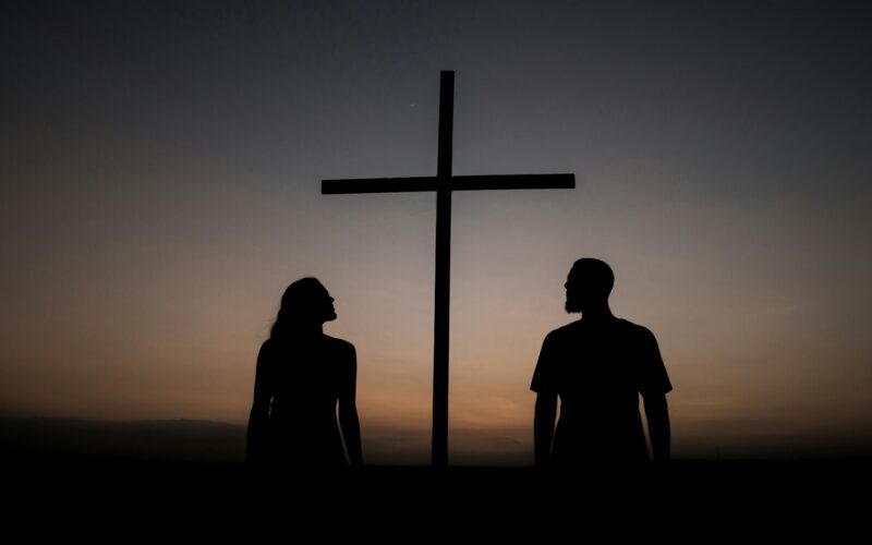 silhouette of man and woman standing beside cross during sunset