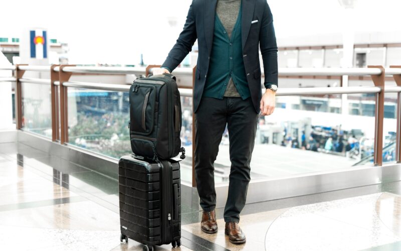 man in blue suit jacket and black pants standing beside black luggage bag