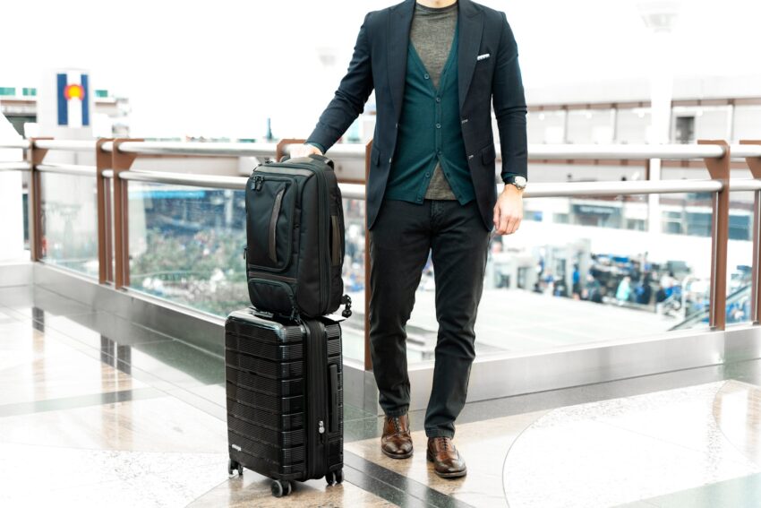 man in blue suit jacket and black pants standing beside black luggage bag